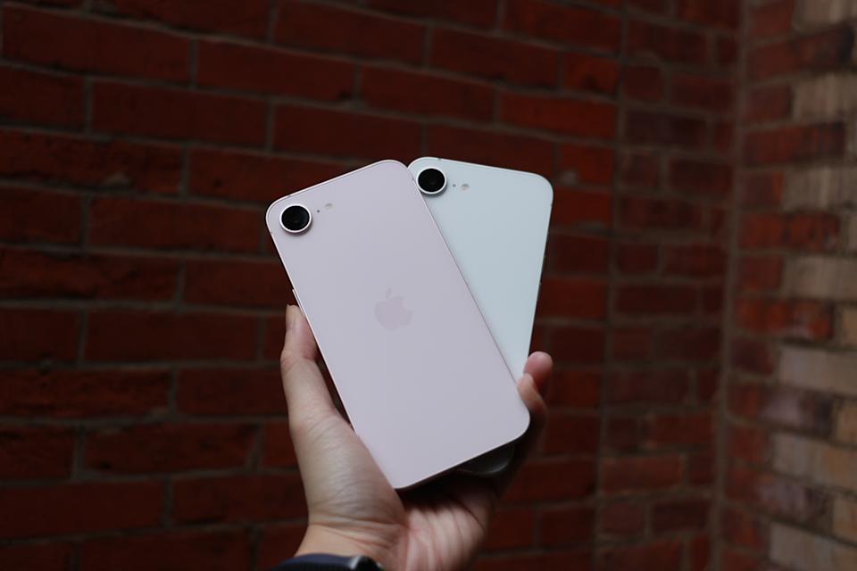 The pink iPhone 17e and the white iPhone 16e held up in one palm in front of a red brick wall.