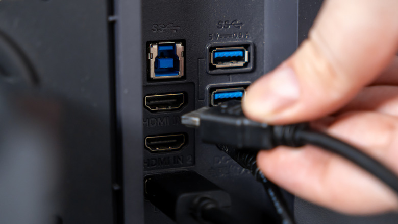A close-up of a monitor's rear ports shows a blue USB-B upstream port, two blue USB-A ports and two HDMI inputs while a cable is being connected.